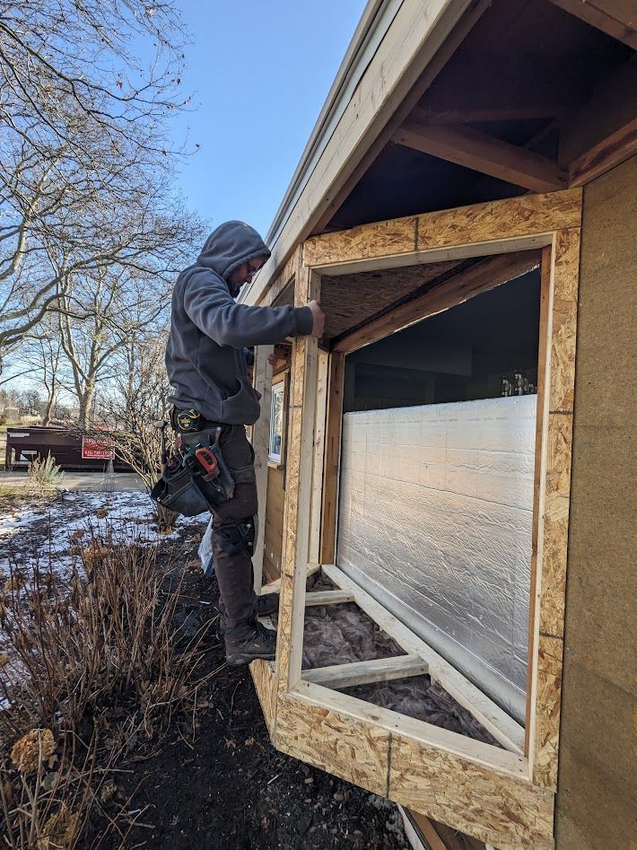 Person in a hooded sweatshirt installing a window frame on a building's exterior.