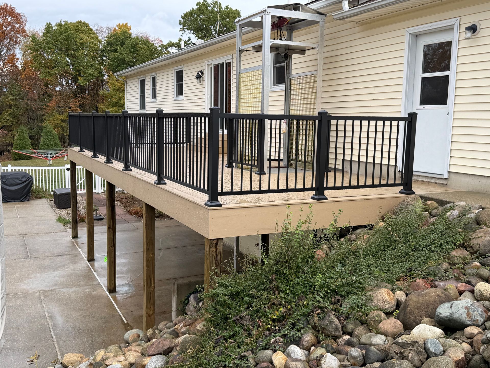 Deck with black railing next to a house with a white door. Landscaping and a wet walkway are in view.