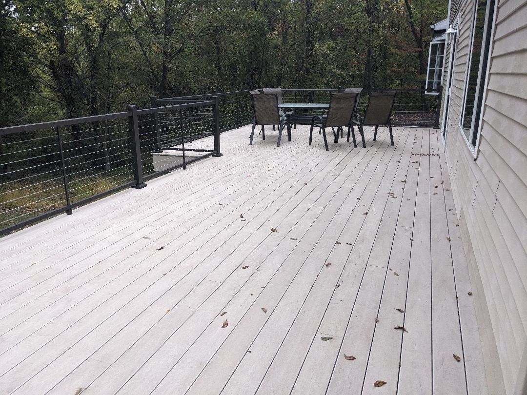 Wooden deck with table and chairs, black railing, trees in background, next to a house.