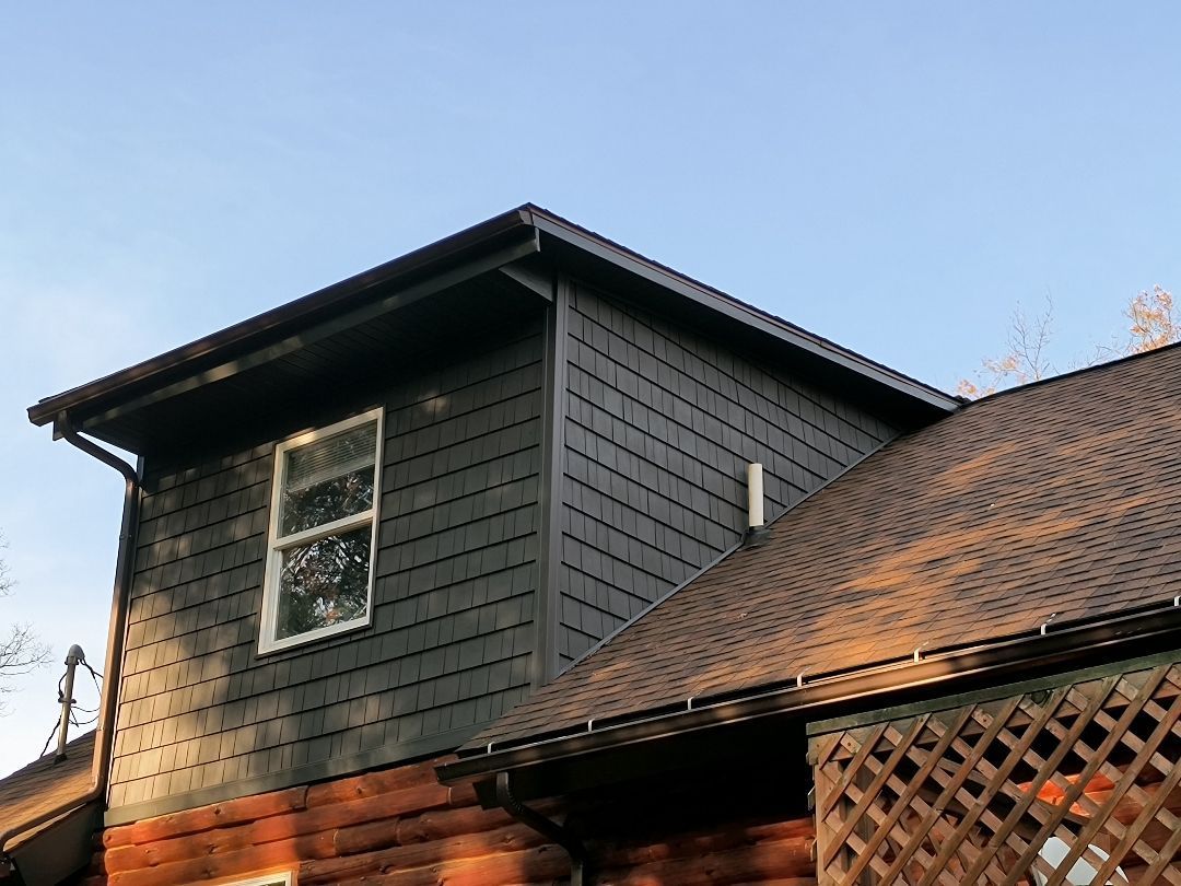 Dark-shingled home addition with a window against a blue sky, connected to a log cabin structure.
