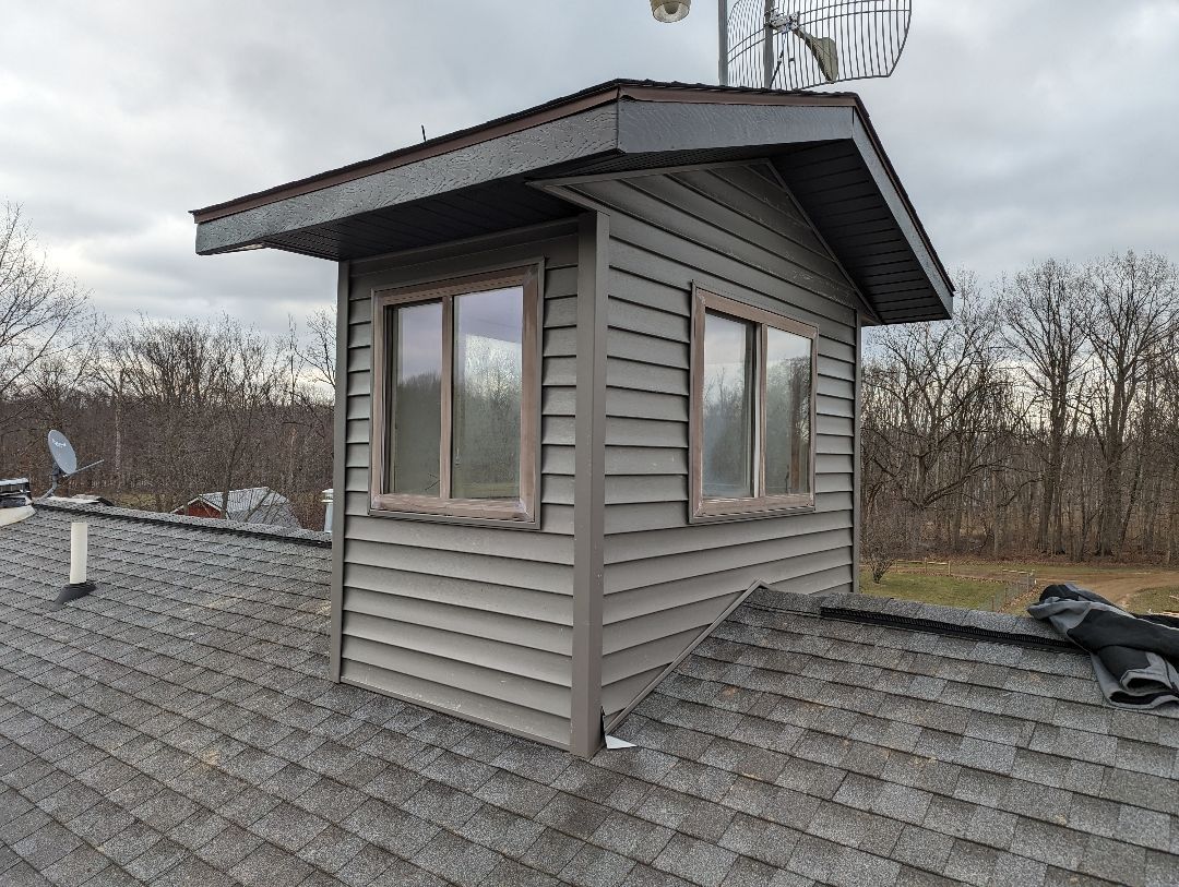 Grey sided building on a dark shingle roof with two windows; a small tower with antenna behind it.