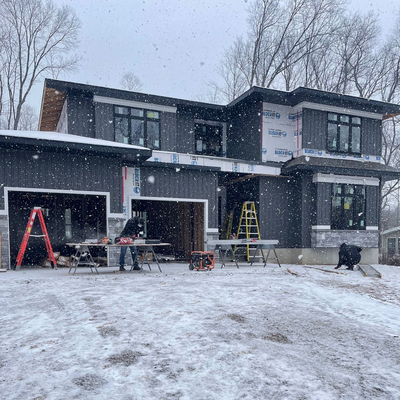 Construction site: two-story modern house with dark siding under snowfall. Workers near tools and garage.