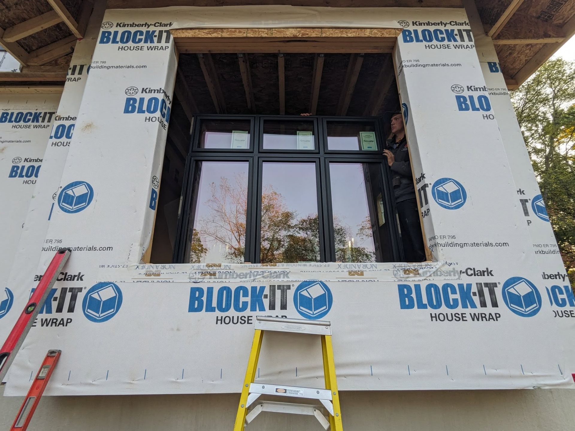 Man installing black window in new house; blue house wrap, yellow ladder, red level.