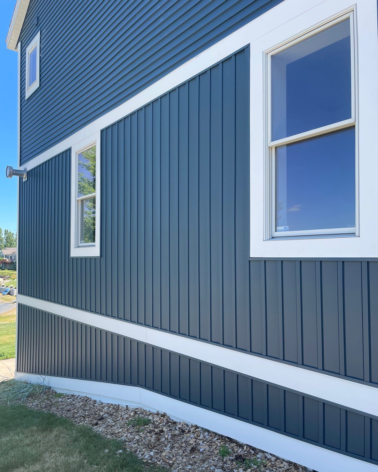Blue siding on a house with white trim and windows, set against a blue sky and green grass.