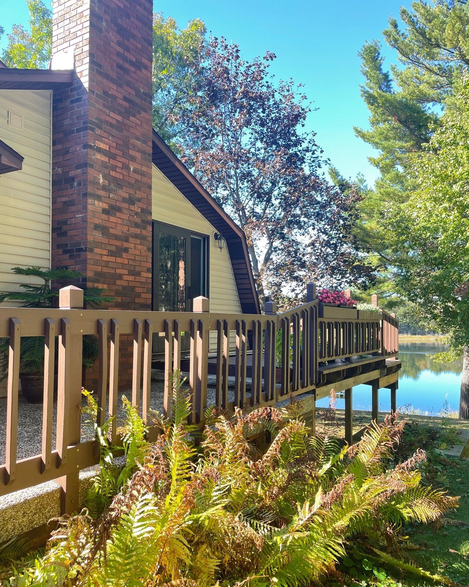 Wooden deck with railing, overlooking a lake, connected to a house with a brick chimney.