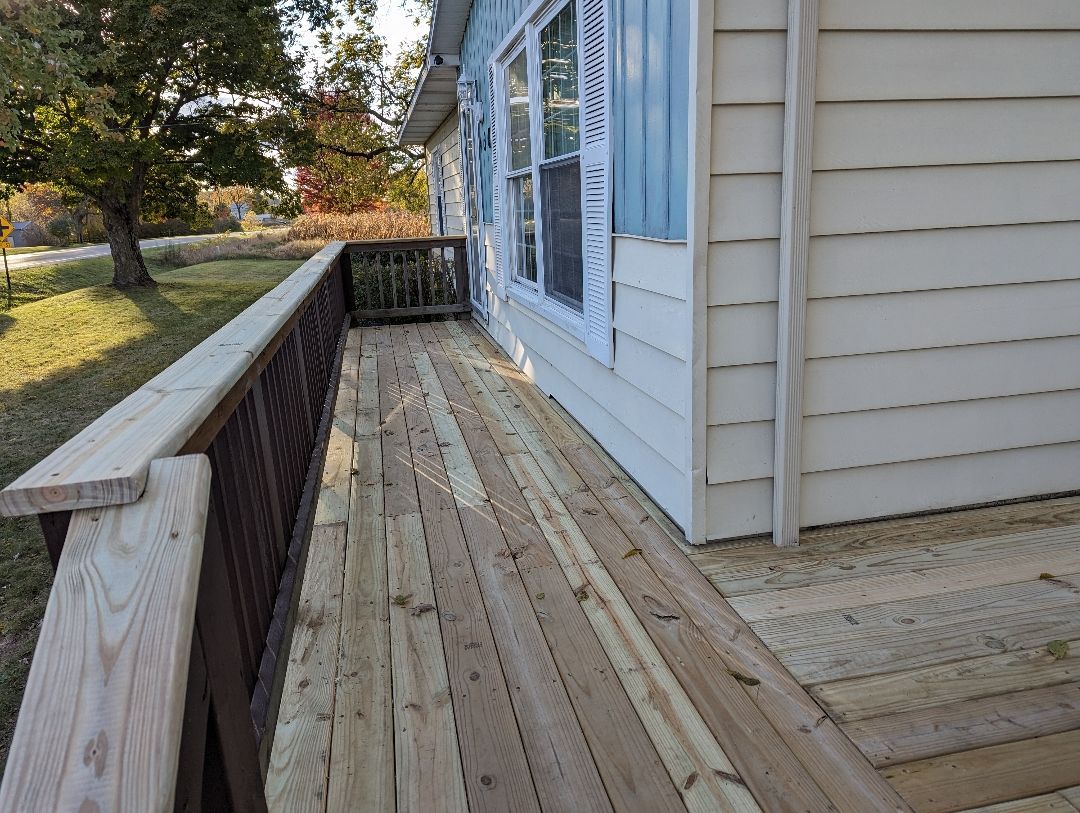 Wooden deck of a house with brown railing, light-colored siding, and two windows.