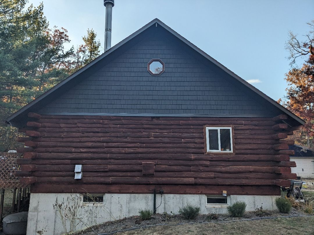 Rustic cabin with log siding, gray shingle roof, chimney, and small round window. A white window on the logs.
