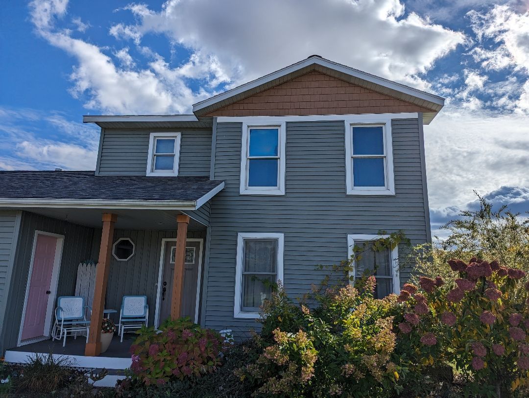 Two-story gray house with white window frames under a cloudy sky. Pink door, bushes in front.