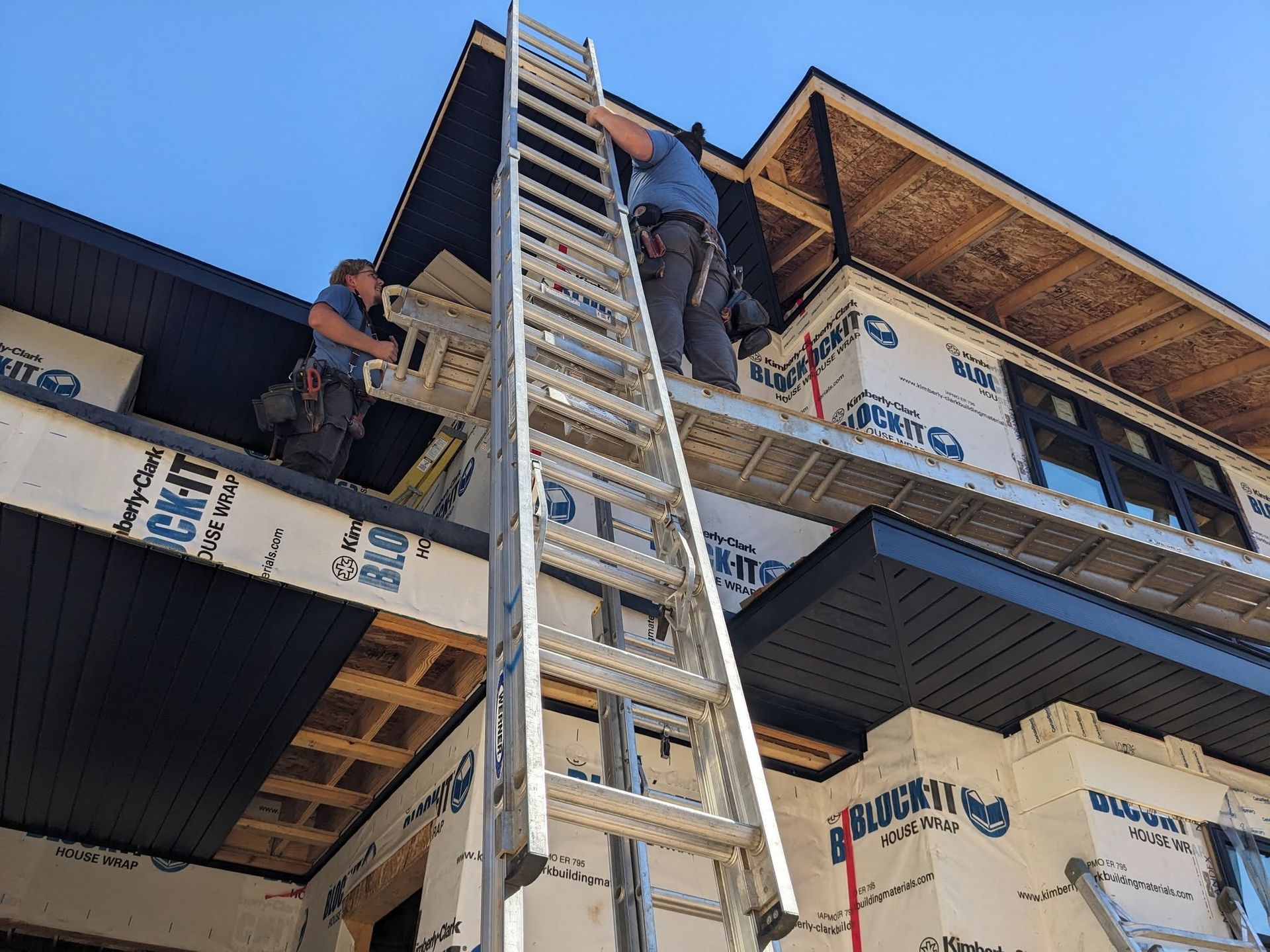 Two construction workers on a roof, using a ladder, installing siding on a building under a blue sky.