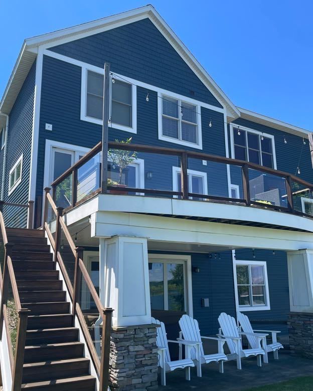 Blue house with white trim and glass railing, wooden stairs, and white Adirondack chairs.