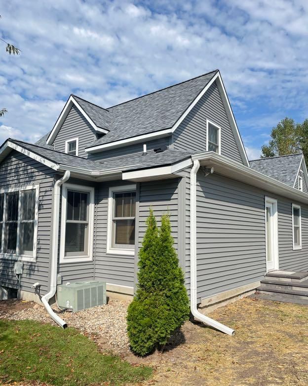 Gray house with gray siding, roof, and white trim, a small evergreen tree, and blue sky.