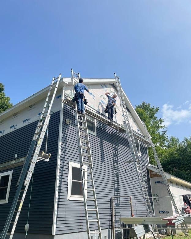 Two workers on ladders installing siding on a house under a blue sky.