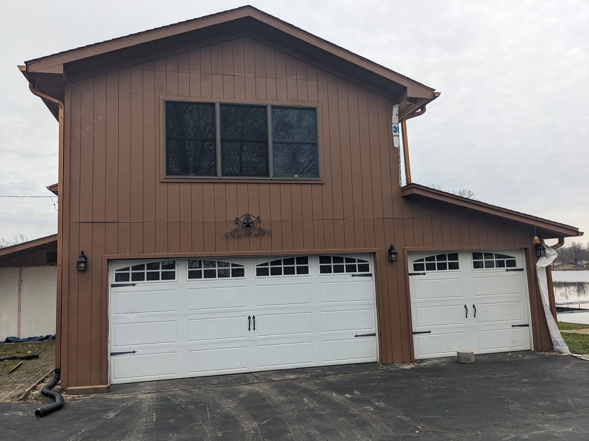 Brown two-story house with white garage doors; overcast sky.