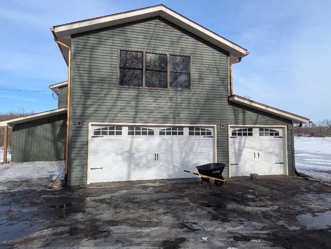 Two-story green garage with white garage doors, snow on the ground, and a wheelbarrow in front.