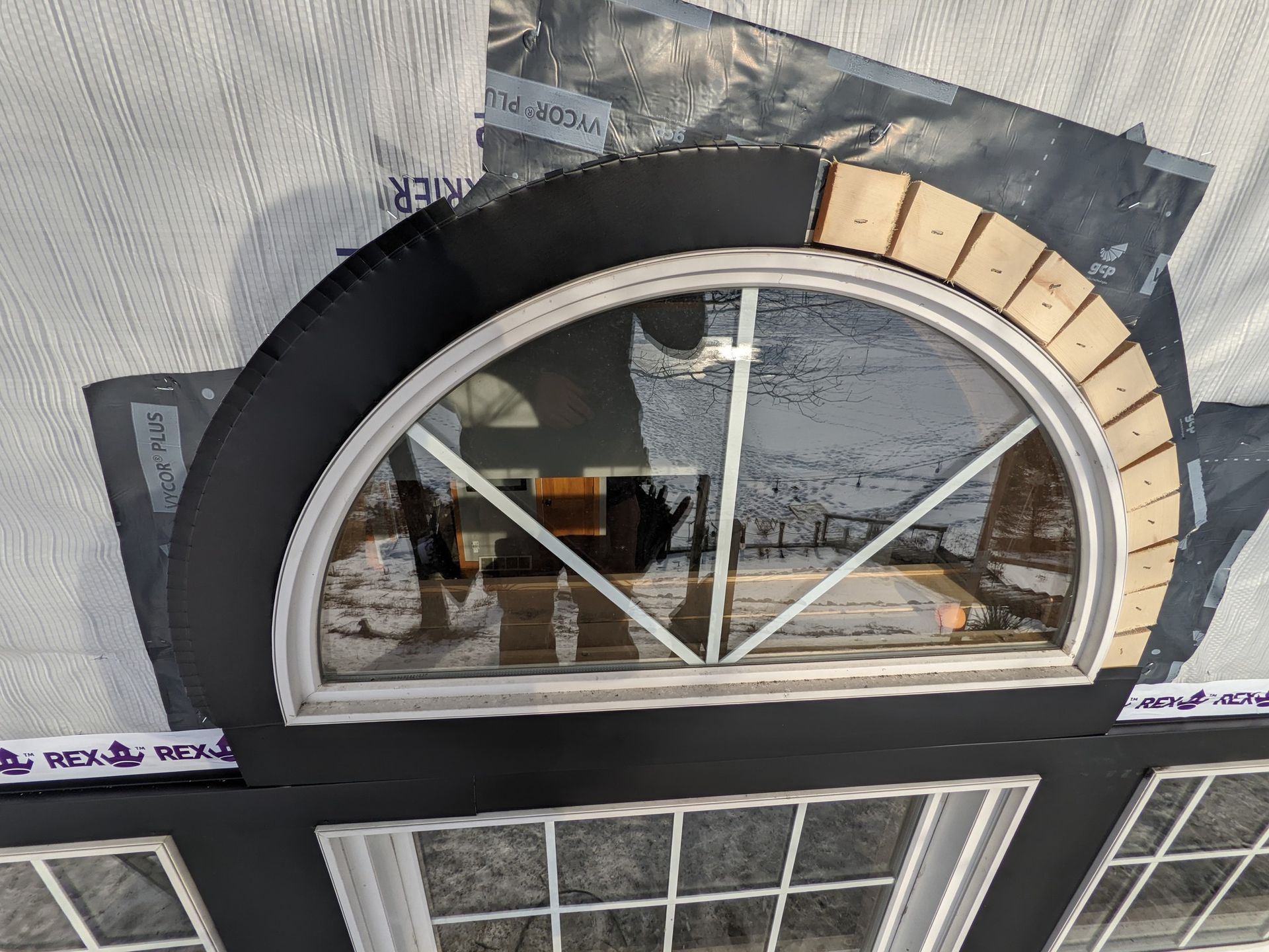 Arched window being installed, with black trim and beige brick veneer, set against a gray tarp.