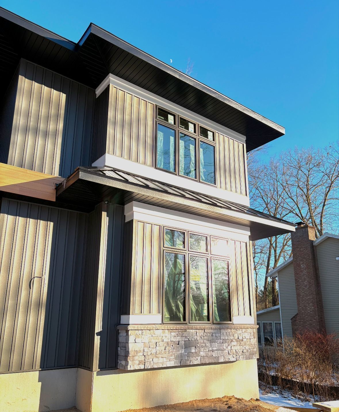Modern house exterior with dark gray siding, large windows, and a stone accent, blue sky.