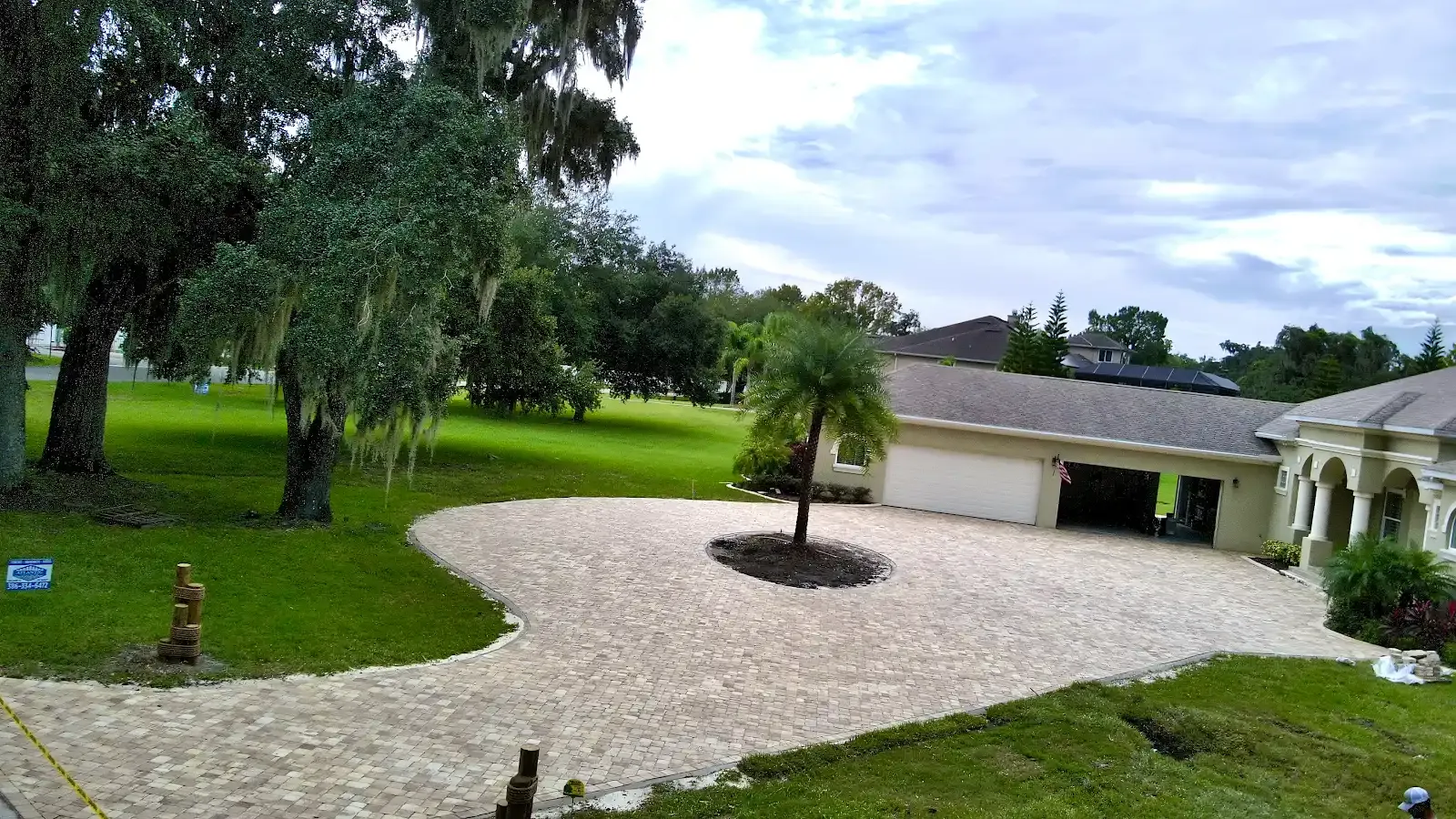 Gravel driveway leading to a garage, surrounded by green grass, trees, and a cloudy sky.