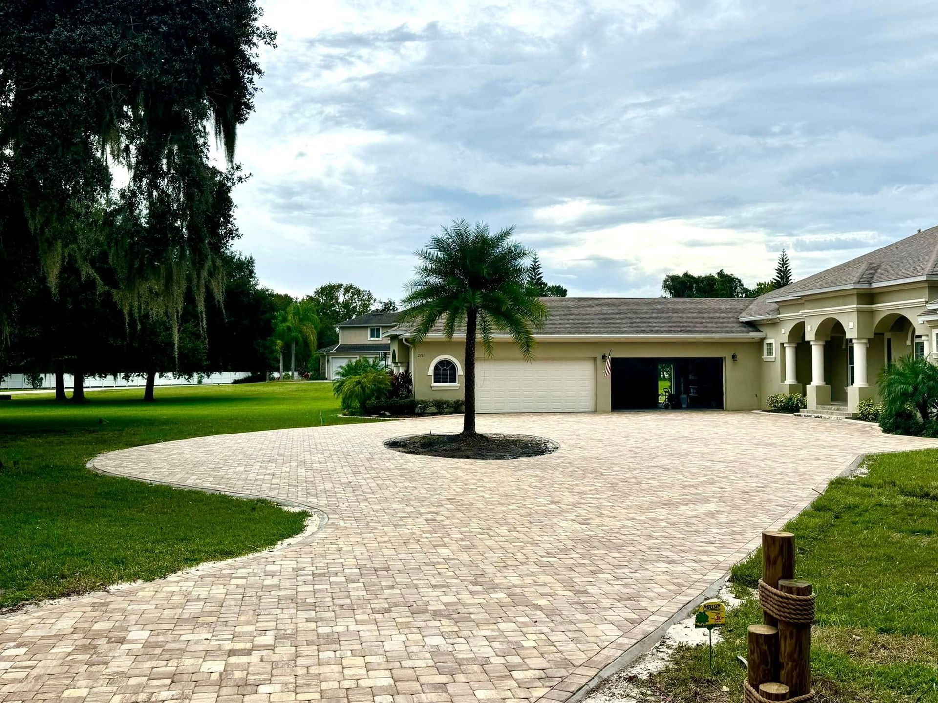 Driveway leading to a beige house and garage with palm tree in the center. Overcast sky.