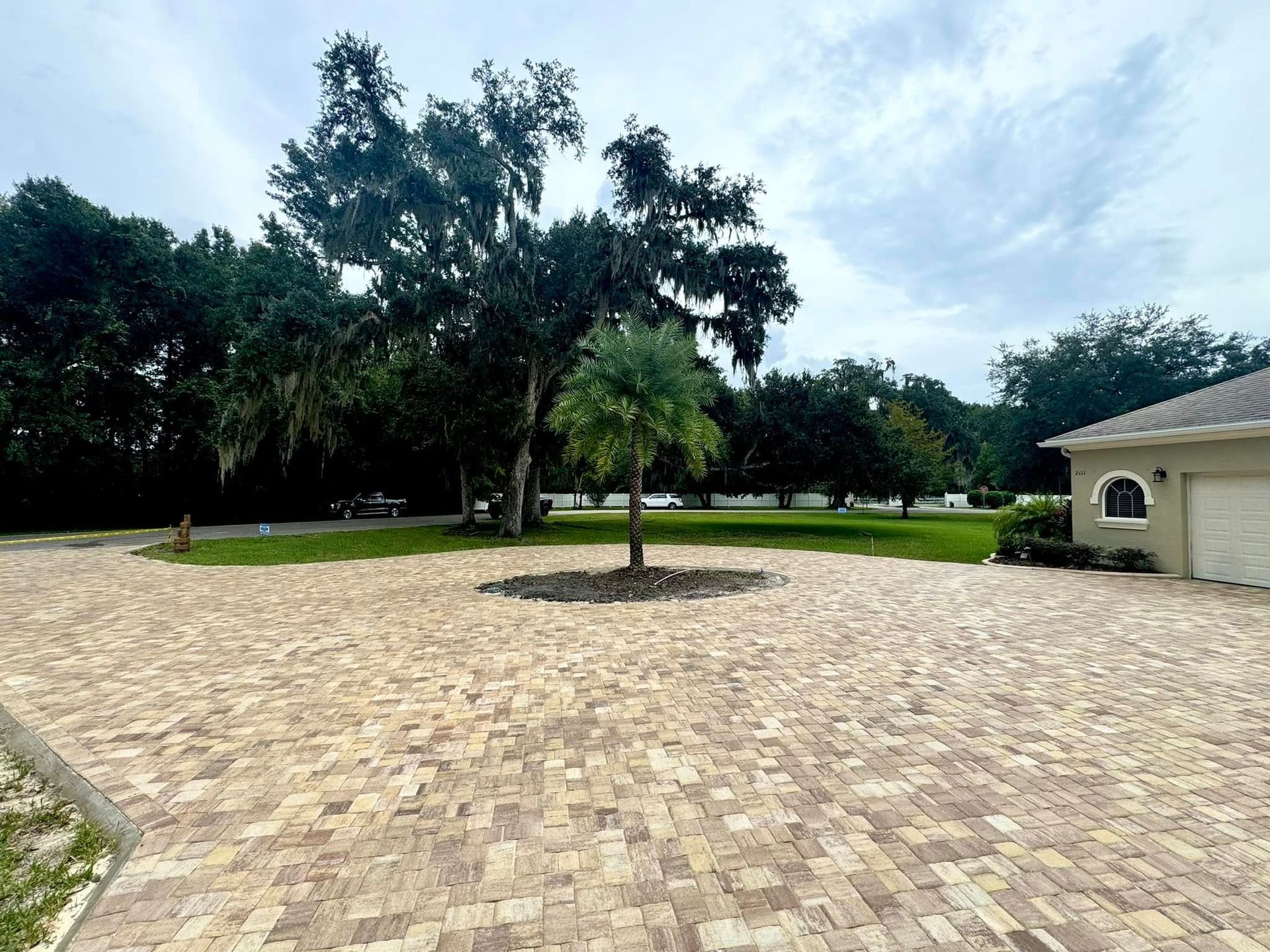 Paver driveway with a tree in the center, and a house on the right, under a cloudy sky.