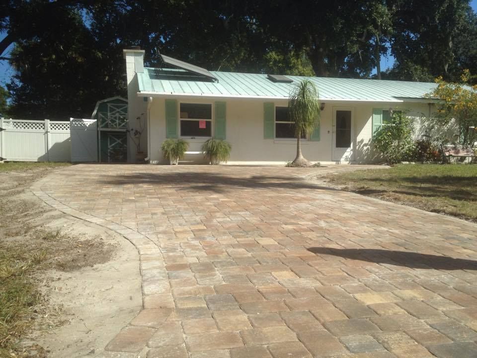 Brick paved driveway leads to a white house with a green roof, shutters, and a small tree in front.