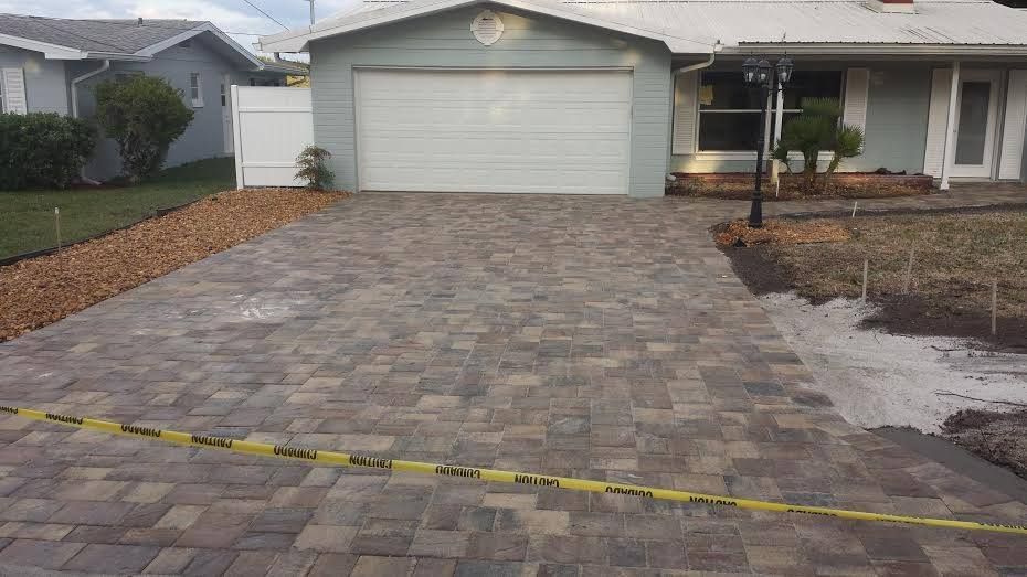 Paver driveway in front of a light blue garage and white house; caution tape in foreground.