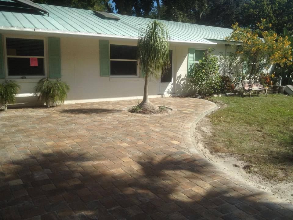 Brick walkway leading to a house with a green roof and shutters. Palm tree in the center, sunny day.