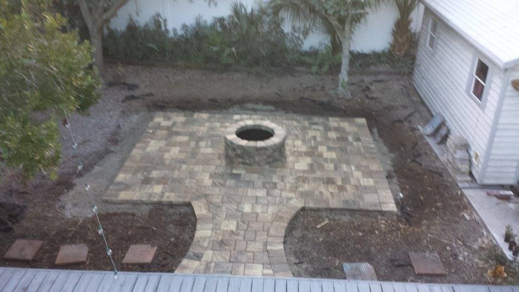 Patio with brick pavers surrounding a stone fire pit in a backyard setting.