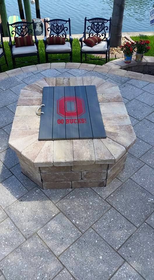 Fire pit with Ohio State logo cover on a brick patio by water.