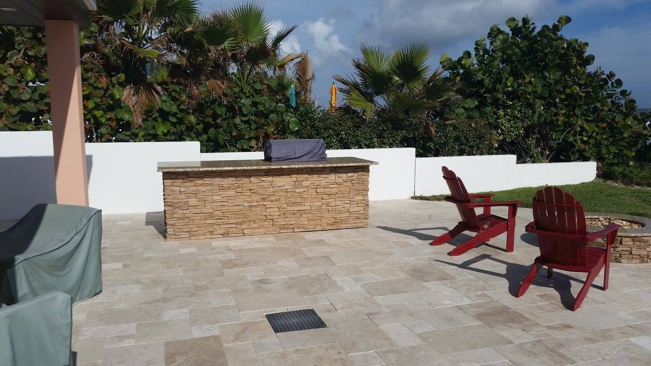 Patio with built-in grill, two red chairs, stone tile, white wall, and lush greenery under a sunny sky.