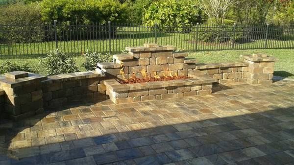Stone fire pit with flames on a paved patio in a yard, backed by a fence and greenery.