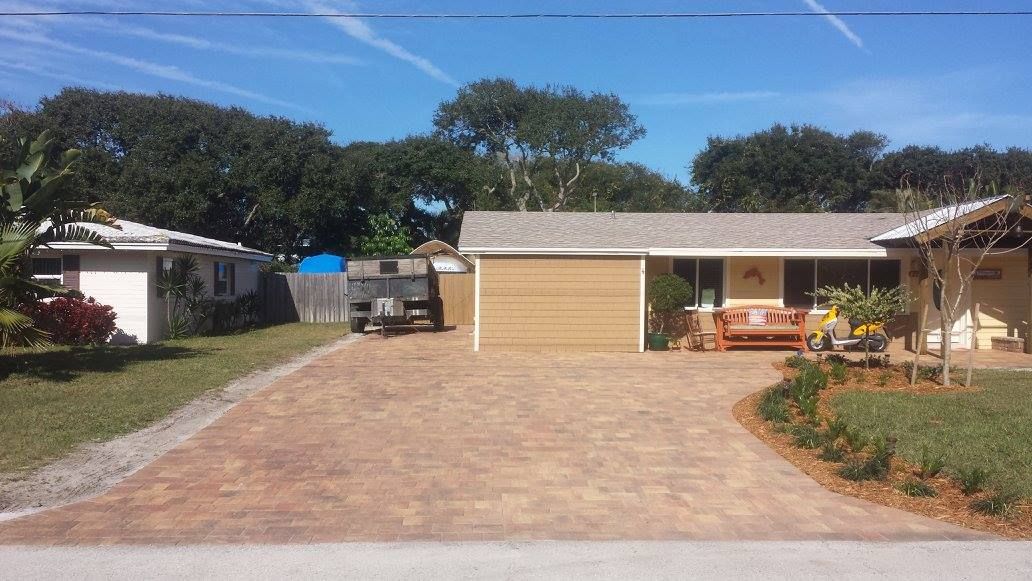 A one-story home with a tan garage, brick driveway, and a white shed on the left.