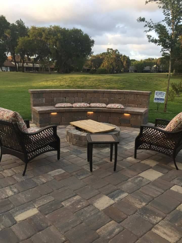 Patio with seating around a fire pit, brick wall with cushions, set on a stone paved area, green grass background.