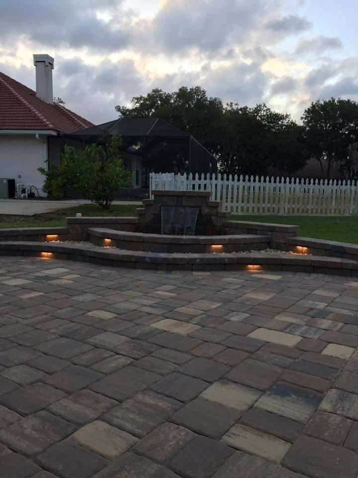 Outdoor patio with a fountain, lit steps, and a white fence. Sky is overcast.