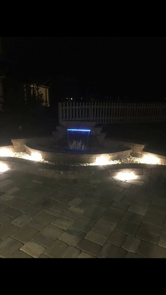 Lit fountain at night with blue light, surrounded by stone patio and a white fence in the background.