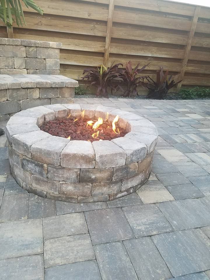 A round stone fire pit with flames, on a patio, with a wooden fence background.