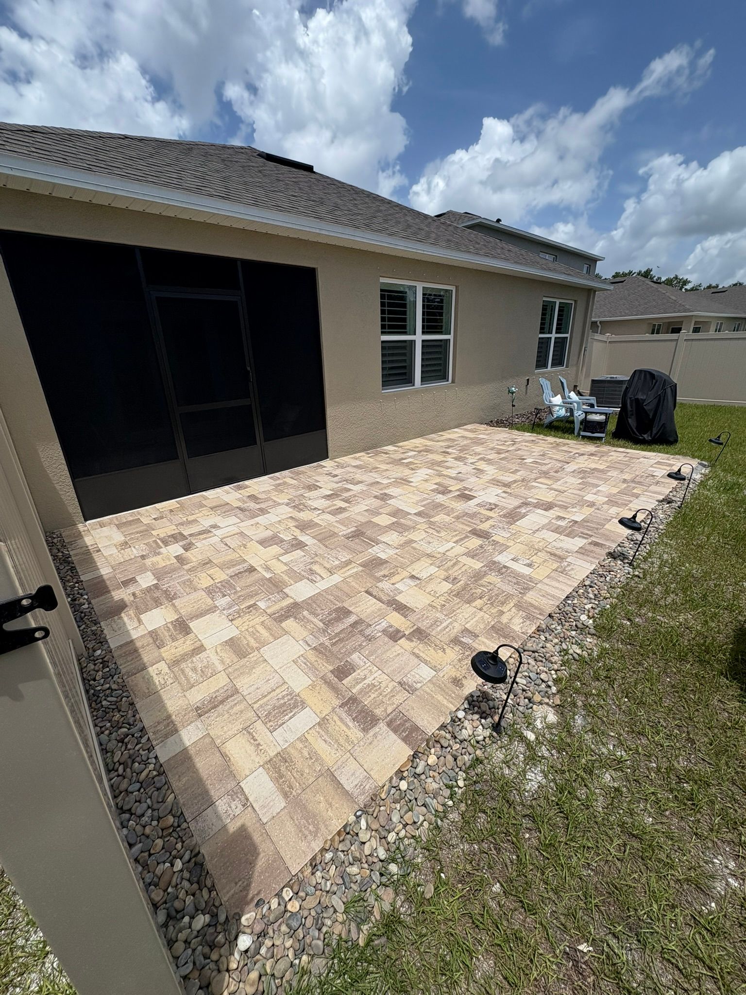 Brick patio adjacent to a beige house, with grass and rocks surrounding the edge.