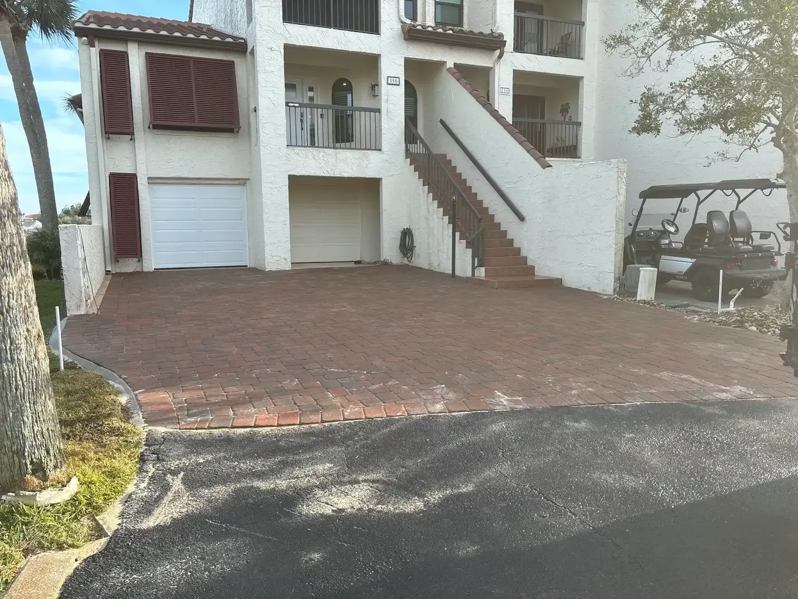 Brick paved driveway leading to a two-story building with a garage, stairs, and parking.
