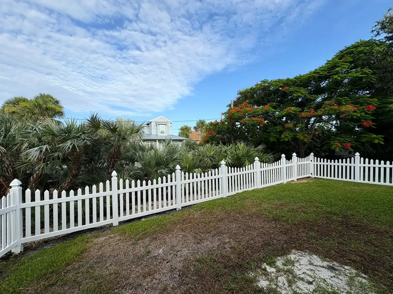 White picket fence in front of a green lawn and trees, with a building in the background against a blue sky.