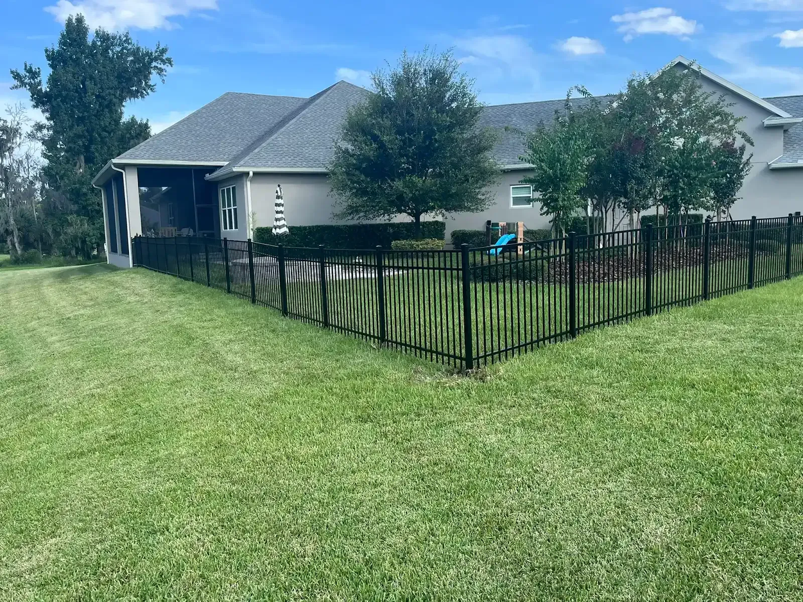 Black metal fence surrounds a grassy yard, partially encircling a light-colored house with a dark roof.