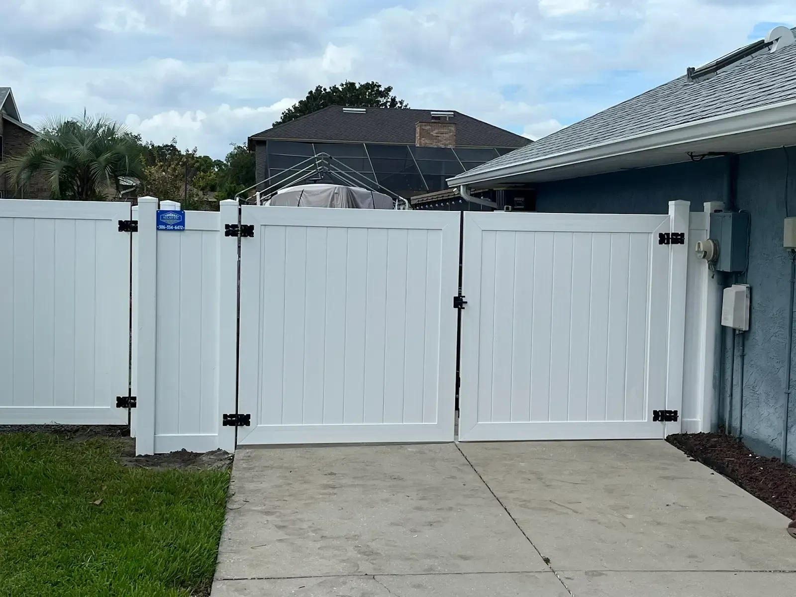 White vinyl fence and double gate on a driveway, black hinges and latch, blue sky.