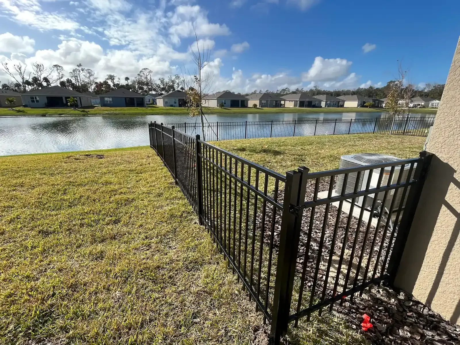 Black fence separating a grassy yard from a lake, with houses in the background under a blue sky.
