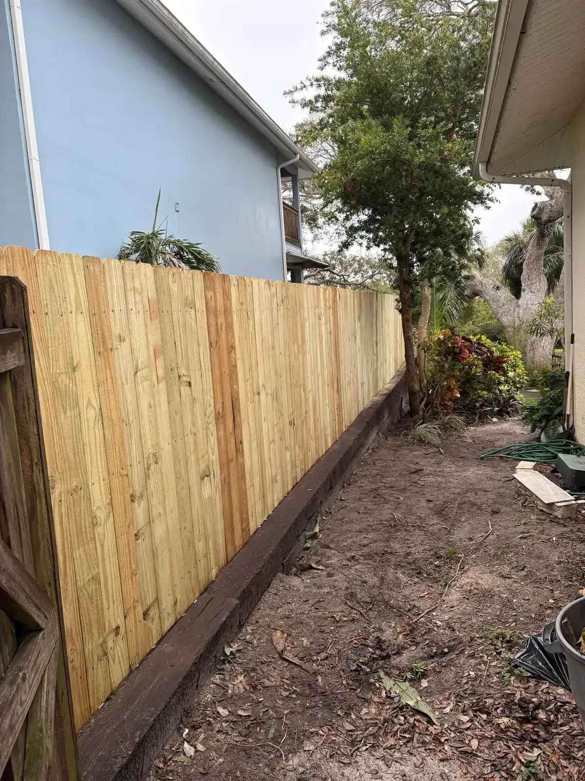 Wooden fence in backyard, next to light blue house and tree.