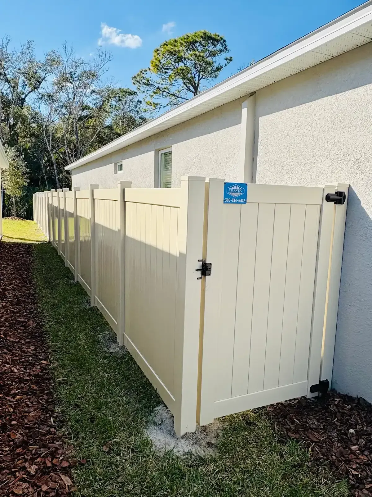 Beige vinyl fence with gate alongside a stucco building, on a sunny day.