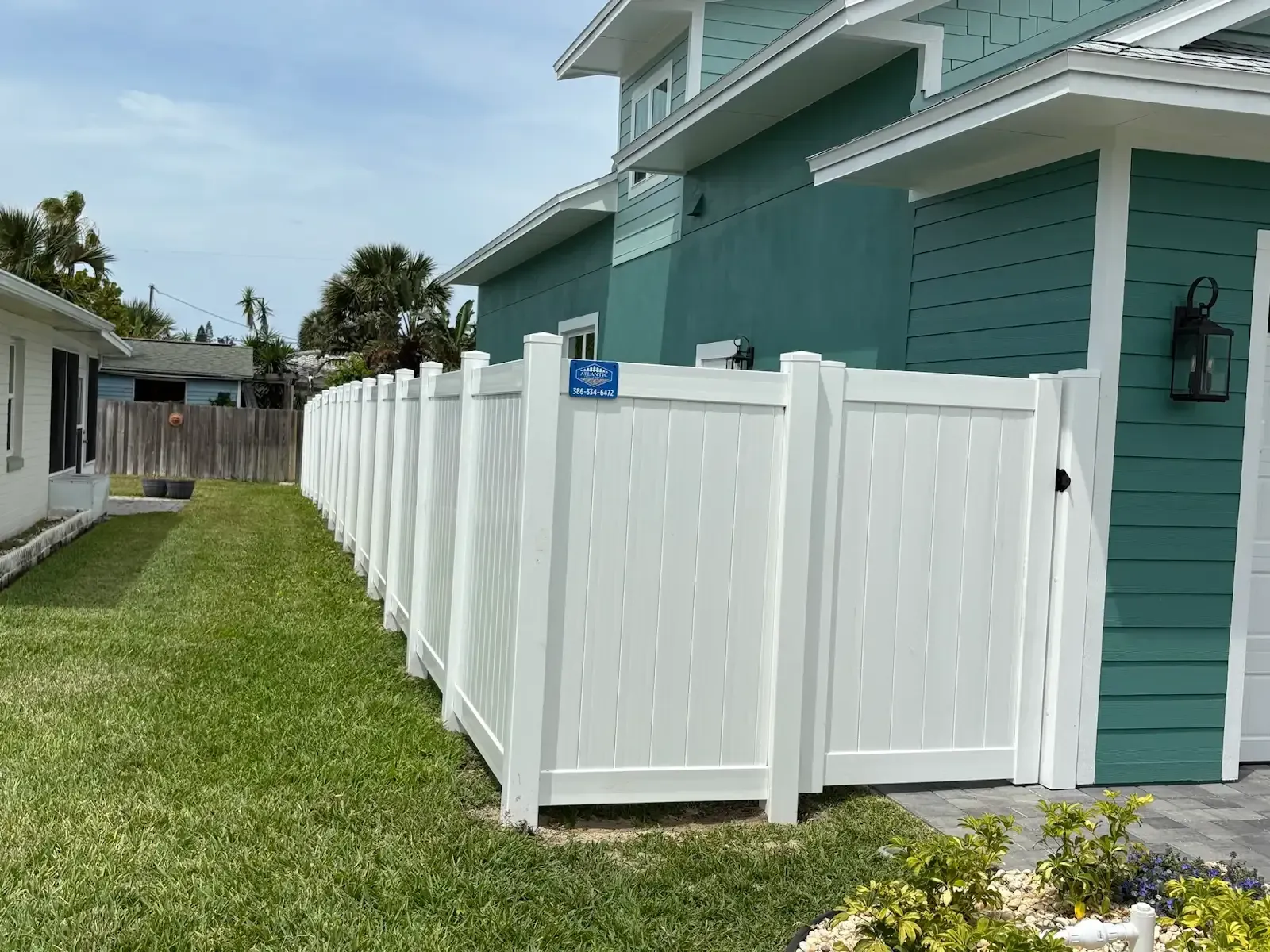 White vinyl fence beside a teal house and green grass on a sunny day.