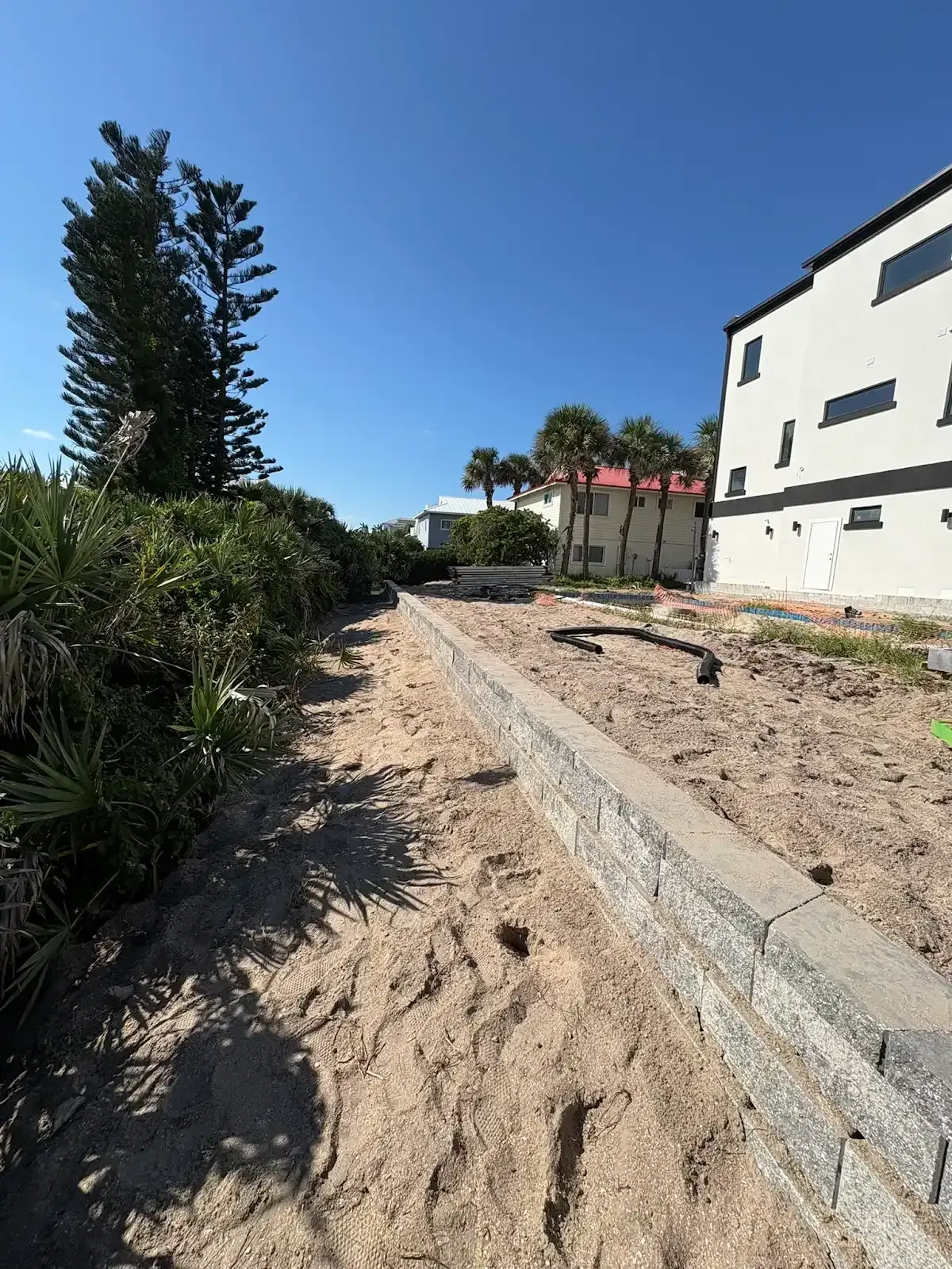 Sandy path next to a low retaining wall, with bushes on one side and a building on the other, under a blue sky.