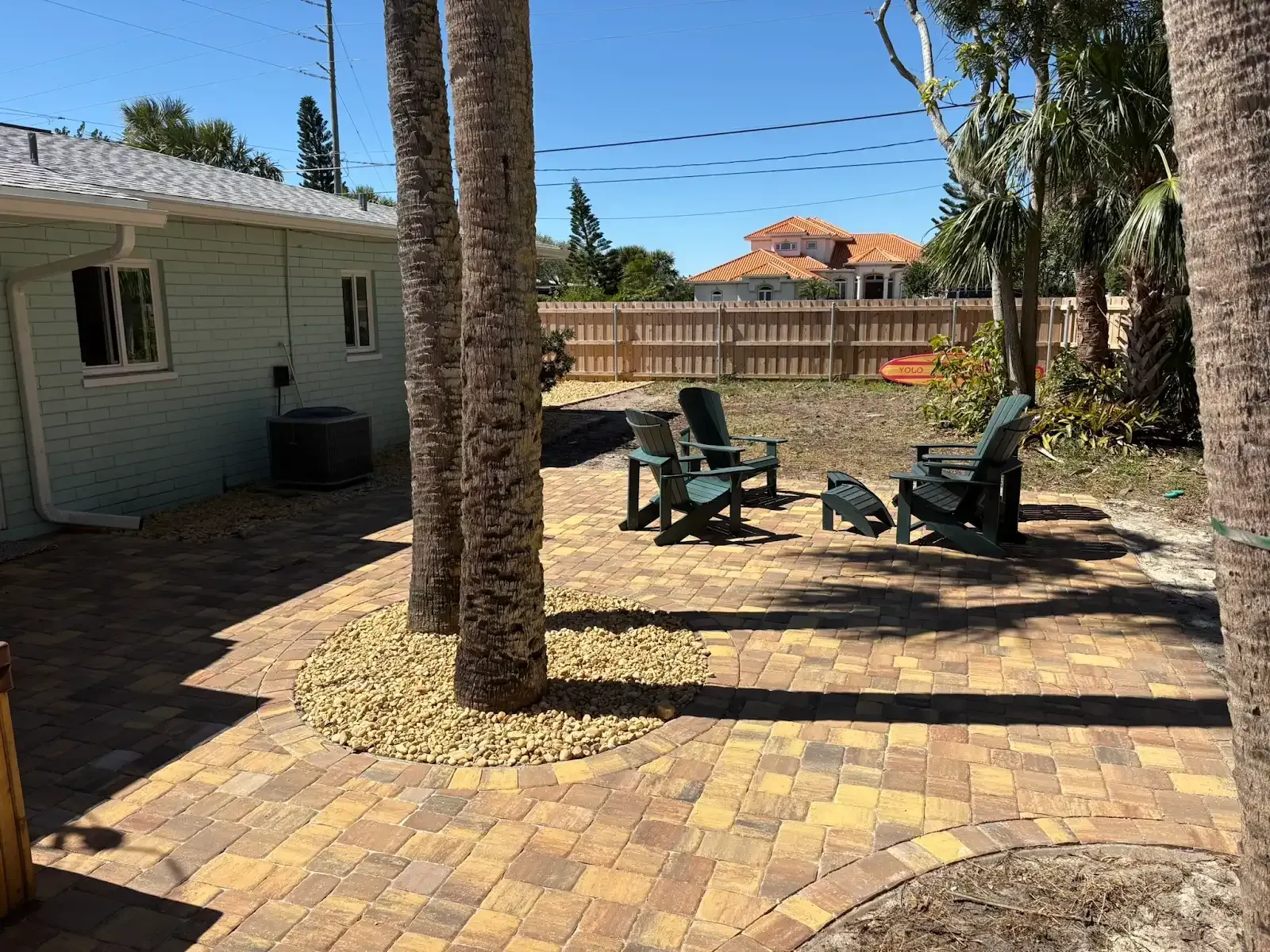 A brick patio with chairs, surrounded by trees and a fence, with a house in the background.