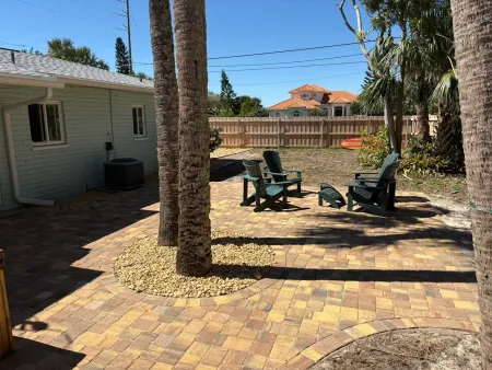 Yellow house with a small garden, palm tree, and a wooden structure in front. Sunny day.