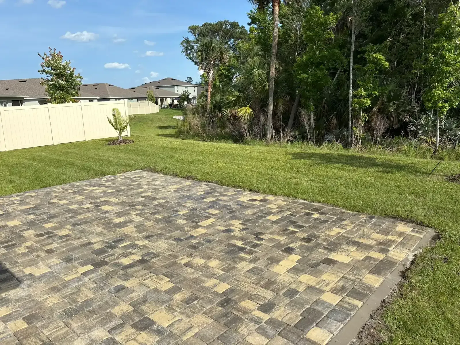 A brick patio in a backyard with green grass, a fence, and trees under a blue sky.
