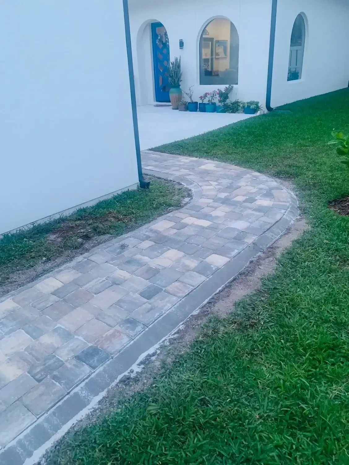 Brick walkway curves through green grass toward a white building with blue door.