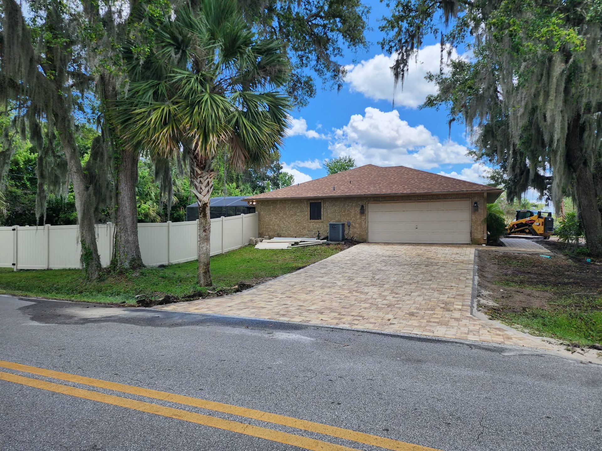 Brick house with attached garage, beige driveway, white fence, and large trees on a sunny day.